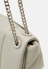 Sac à main en cuir synthétique beige avec une finition texturée, présentant une surface mate et une bandoulière chaîne argentée avec détails œillets.