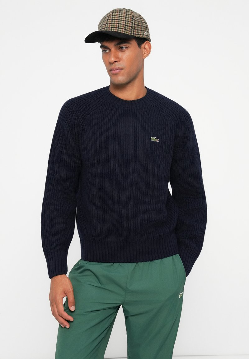 Marinblå ribbad stickad tröja med långa ärmar, rundad halsringning och grön logotyp, i kombination med gröna byxor och en rutig keps.