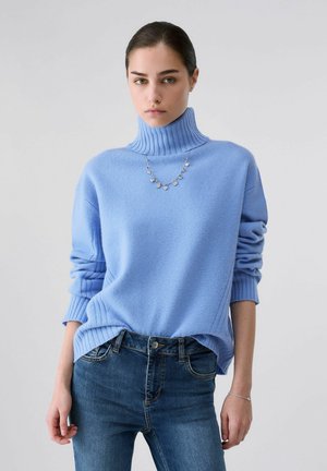 Giovane donna che indossa un maglione a collo alto azzurro chiaro, jeans blu, una collana d'argento e un braccialetto d'argento su uno sfondo chiaro uniforme.
