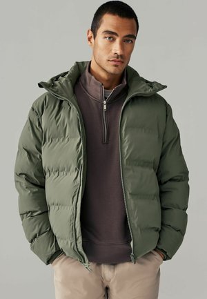 Puffer jacket in olive green, zip-front design with a hood, featuring padded texture and elastic cuffs, layered over a dark gray top.