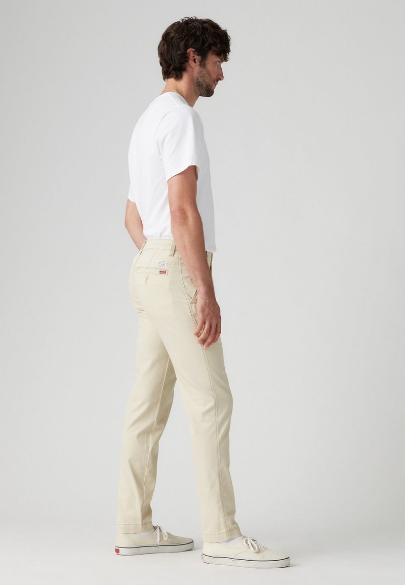 Pantalones de algodón beige con un diseño de pierna recta, bolsillos traseros y detalles de costura sutiles. Combinados con una camiseta blanca lisa y zapatos.
