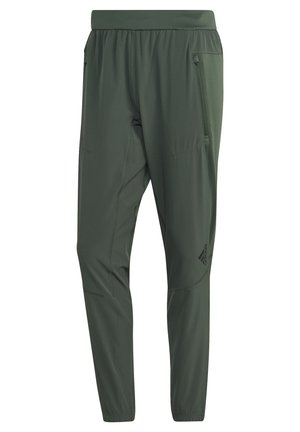 Persona con camiseta sin mangas negra, pantalones verdes y zapatillas negras saltando la cuerda sobre fondo blanco.