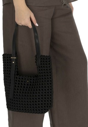 Ręka trzyma czarną plecioną torebkę z paskami ze skóry, nosi brązowe spodnie o szerokich nogawkach.