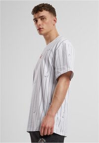 Bílé tričko s krátkým rukávem a vertikálními černými pruhy, s malým červeným logem. Má volný střih a je vyrobeno z měkké bavlny.