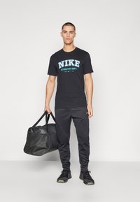 Juodas Nike marškinėlis su mėlyna logo, juodos sportinės kelnės ir juodi sportiniai batai; laiko juodą kibirą su petnešomis.