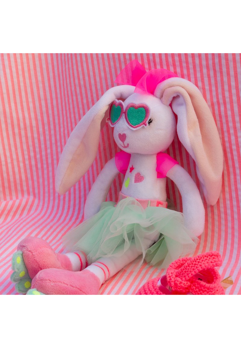 Λούτρινο κουνελάκι με λευκή γούνα, μεγάλα ροζ αυτιά, γυαλιά ηλίου σε σχήμα καρδιάς και ροζ μπλούζα. Φοράει πράσινη φούστα από τούλι και έχει ροζ πόδια.