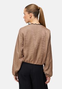 Brun, tekstureret bluse med lange ærmer, elastisk talje og flæsedetalje ved halsudskæringen, kombineret med sorte bukser med høj talje.