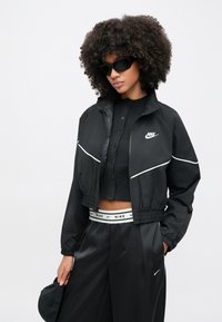 Μαύρο κοντό μπουφάν Nike με λευκές λεπτομέρειες, κλείσιμο με φερμουάρ και ψεύτικο γιακά, σε συνδυασμό με μαύρο ριμπ τοπ και φαρδιά μαύρη παντελόνια.