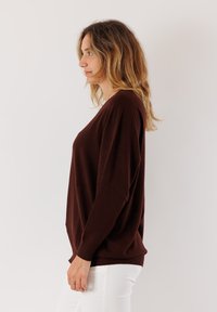 Donna con capelli biondi mossi, che indossa un maglione a maniche lunghe marrone scuro e pantaloni bianchi, in posizione di profilo su uno sfondo semplice.