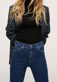 Mujer rubia vestida con un blazer de cuadros oscuros, un jersey de cuello alto negro y unos jeans de tiro alto azules, con las manos detrás de la espalda.