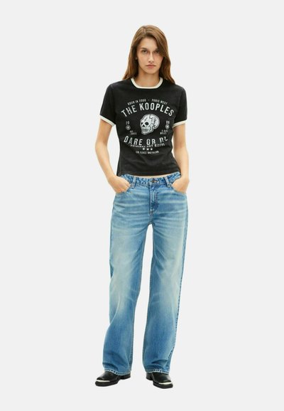 Juoda marškinėliai su baltu kaukolės piešiniu ir tekstu, derinami su šviesiai mėlynomis plačiomis kelnėmis. Modelis stovi su rankomis kišenėse, dėvėdamas juodus batus.