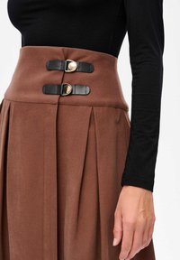 Hooggesneden bruine rok met plooien en twee zwarte leren gespsluitingen, gedragen met een zwart longsleeve-hemel, waarbij de rechterarm en hand zichtbaar zijn.