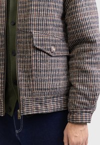 Veste en mélange de laine avec un motif pied-de-poule en gris, marron et blanc. Elle dispose d'une fermeture éclair, de poches latérales et de poignets côtelés.