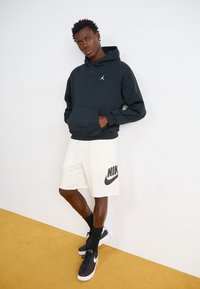 Svart huvtröja med en känguruficka och Jordan-logotyp, ihopsatt med gräddfärgade shorts med en svart Nike-logotyp. Svarta strumpor och sneakers fulländar looken.