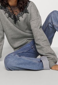 Šedá mikina s černým krajkovým výstřihem a volným střihem, spárovaná s modrými džíny s ohrnutými manžetami. Měkký materiál s výrazným kontrastem textur.