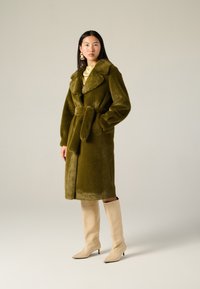 Cappotto in pelliccia finta verde oliva con ampi risvolti, cintura in vita e lunghezza fino al ginocchio; abbinato a stivali con tacco beige alti fino al ginocchio.