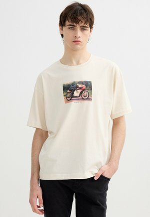 Joven con camiseta crema con un gráfico de una moto de carreras vintage con el número 15, combinada con pantalones negros.