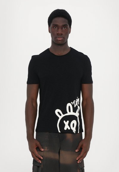 T-shirt en coton noir avec un col rond et des manches courtes, arborant un graphique de style graffiti blanc sur le bas devant et sur le côté.