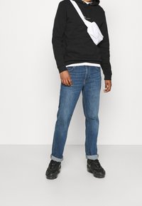 Sudadera negra con bolsillo frontal, vaqueros de denim azules y zapatillas negras. Un bolso cruzado blanco añade contraste al conjunto.