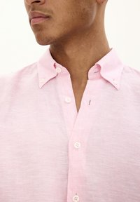 Camicia di lino rosa chiaro con colletto button-down, caratterizzata da bottoni bianchi e un piccolo dettaglio di cucitura vicino al collo.