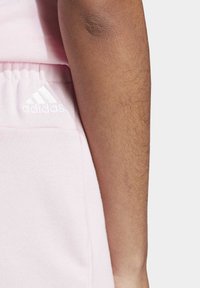 Svetlo roza Adidas trenirke z elastičnim pasom, ki imajo belo logo na levi strani in mehak, teksturiran material.