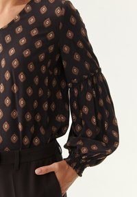 Schwarze Bluse mit braunen geometrischen Mustern, puffärmeliger Schnitt mit gerafften Manschetten und V-Ausschnitt, aus einem leichten Stoff gefertigt.