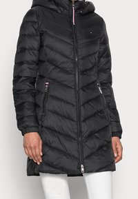 Donna che indossa un lungo cappotto invernale nero trapuntato con zip frontale, tasche con zip, polsini elasticizzati e cappuccio, abbinato a pantaloni bianchi.
