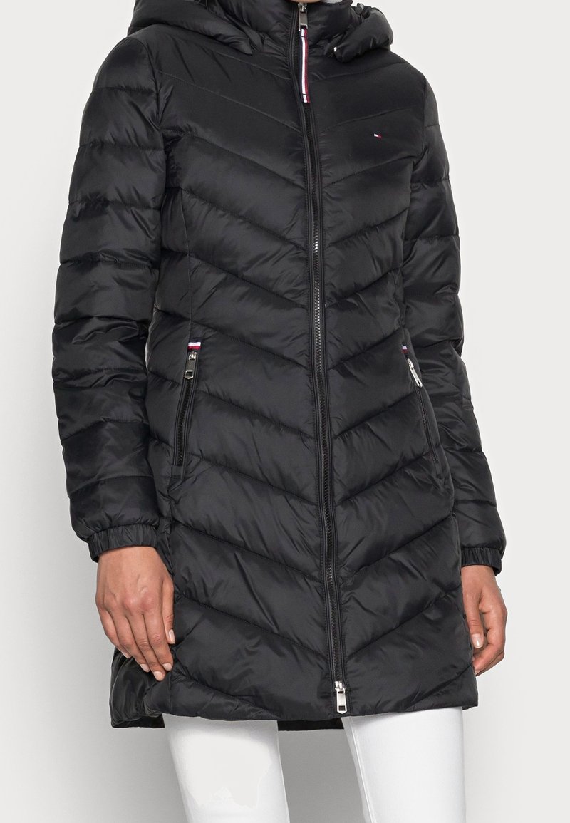 Donna che indossa un lungo cappotto invernale nero trapuntato con zip frontale, tasche con zip, polsini elasticizzati e cappuccio, abbinato a pantaloni bianchi.