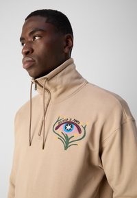 Beige zip-up sweatshirt featuring an embroidered eye design with colorful accents and floral elements on the left chest. Soft fabric texture.