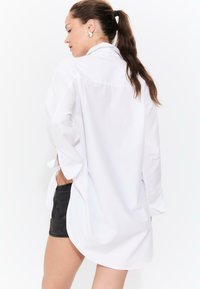 Woman with brown ponytail wearing oversized white button-up shirt and black denim shorts, standing with one hand in pocket, looking left.