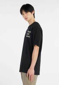 Camiseta de algodón negra con texto blanco en la parte delantera, cuello redondo y corte relajado. Modelo con pantalones beiges, de pie de lado.