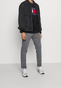 Zwarte spijkerjas over een donker trui met een rode en witte streep. Grijze broek en grijs-witte sneakers maken de outfit compleet.