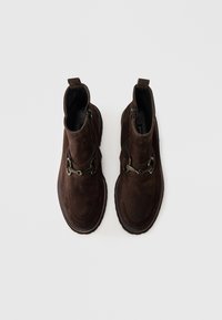Une paire de bottines en daim marron foncé avec des zips latéraux et des détails décoratifs en métal en forme de mors de cheval, photographiée de dessus sur un fond blanc.