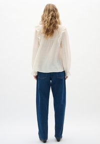 Blusa leggera color crema con dettagli a volant sulle spalle, abbinata a jeans in denim blu scuro a taglio comodo. Tessuto liscio con una sottile trama.