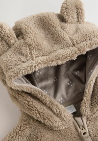 Beige Fleece-Hoodie mit weichem Textil, Bärenohren und gefüttertem Innenraum mit einem subtilen Punktmuster. Zipper-Verschluss vorne.