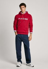 Sudadera de algodón roja con logo blanco, bolsillo frontal y capucha con cordones; combinada con pantalones azul marino y zapatillas grises.