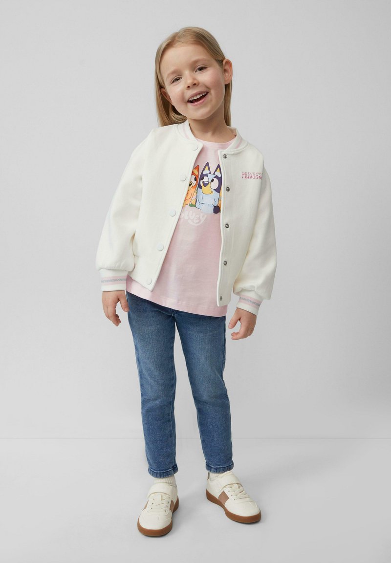 Witte bomberjack met knoopsluiting, roze graphic t-shirt met cartoonfiguren, blauwe jeans en witte sneakers met bruine zolen.