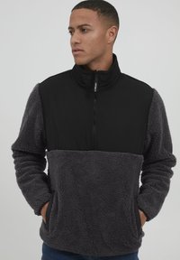 Grå fleece pullover med et sort øverste panel, høj krave, lynlås lukning og sidelommer. Teksturen er blød og hyggelig.