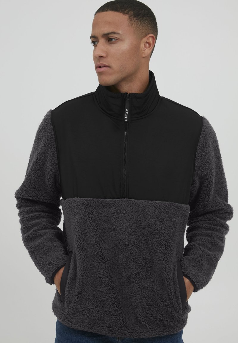 Grå fleece pullover med et sort øverste panel, høj krave, lynlås lukning og sidelommer. Teksturen er blød og hyggelig.
