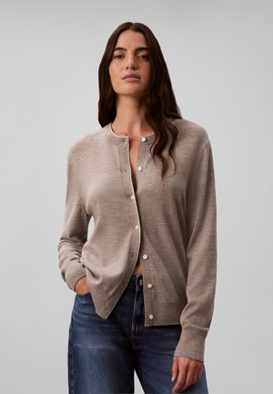 Femme aux longs cheveux foncés, portant un cardigan beige boutonné et un jean bleu, debout avec une main dans la poche devant un fond gris uni.