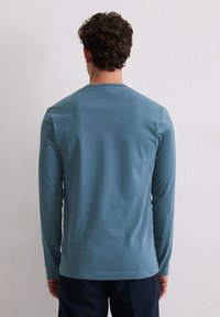 Koszulka z długim rękawem i okrągłym dekoltem, wykonana z miękkiej tkaniny w kolorze mięty. Posiada bezszwowy tył, lekko dopasowany krój oraz elastyczne mankiety.