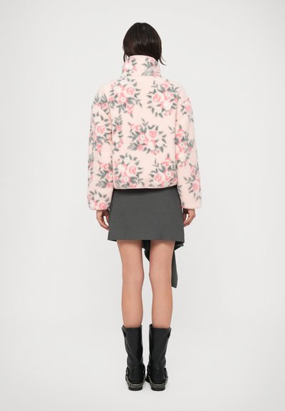 Fleecová bunda svetloružovej farby s kvetinovým vzorom v odtieňoch sivej a ružovej. Má vysoký golier a voľný strih. Kombinovaná s tmavosivou sukňou.