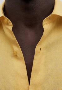 Camisa de lino amarilla con cuello abotonado, que presenta una pequeña muesca y un acabado de tela texturizada. Primer plano de la zona del cuello.