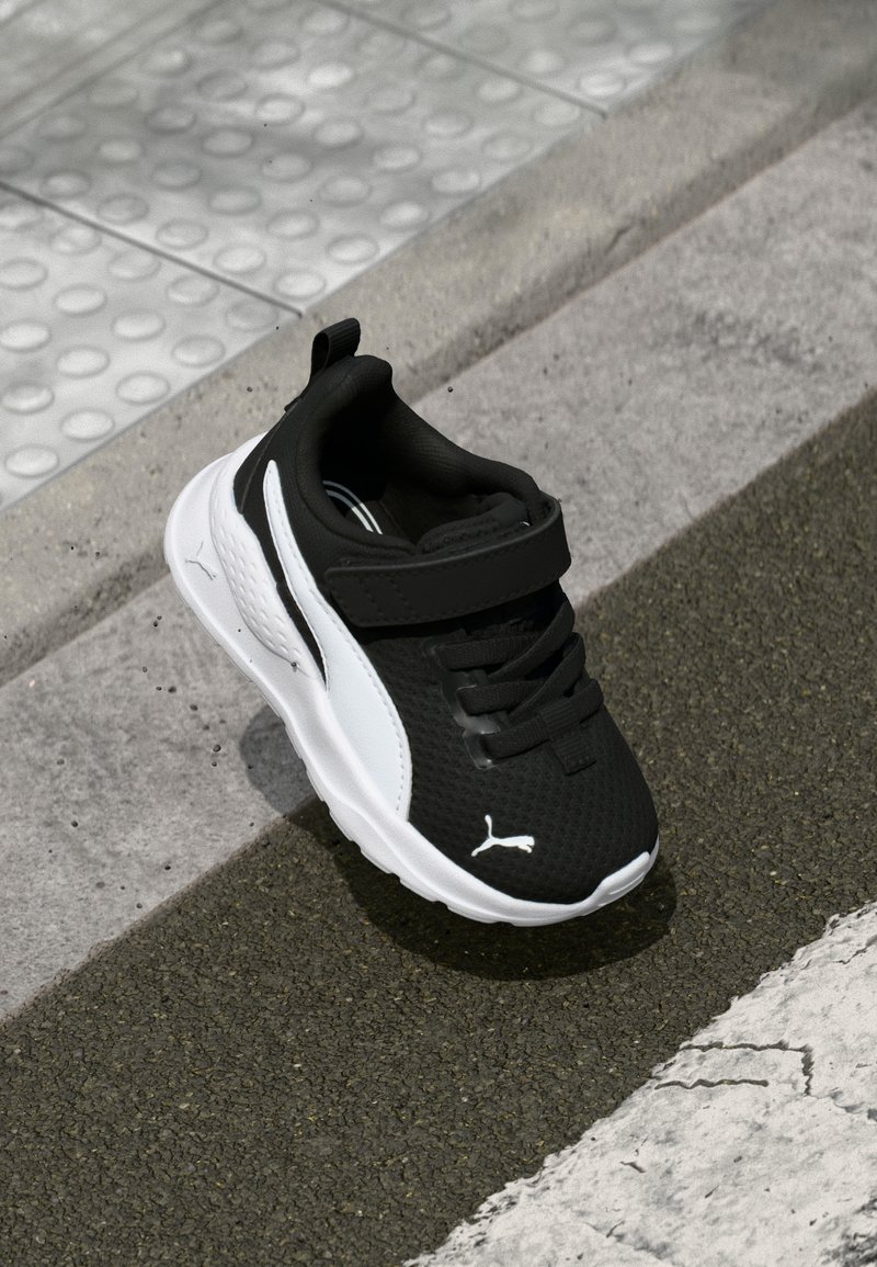 Chaussure de sport noir et blanc avec tige en maille, col rembourré et sangle Velcro, présentant une semelle texturée et un logo sur le côté.