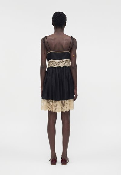 Γυναίκα με πλάτη φοράει μαύρο αμάνικο φόρεμα με μπεζ δαντέλα στη μέση και στο τελείωμα, συνδυασμένο με σκούρα κόκκινα φλατ παπούτσια.