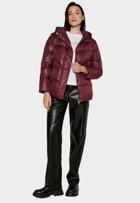 Giacca imbottita color borgogna con cappuccio, texture lucida, chiusura con bottoni a pressione e ampi pannelli trapuntati, abbinata a pantaloni neri in ecopelle e stivali.