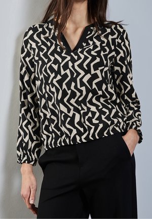 Vrouw die een zwart-wit abstract patroon blouse met lange mouwen met elastische manchetten en een zwarte broek draagt, poseert met één hand in haar zak.