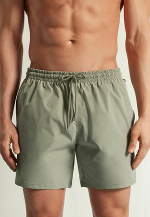 Męski tors w jasnografitowych elastycznych szortach z regulowanym sznurkiem, stojący z rękami swobodnie opuszczonymi wzdłuż ciała na neutralnym tle.