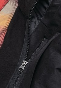 Giacca nera con una cerniera visibile, caratterizzata da una texture liscia e una fodera a quadri in colori caldi. Il tirante della cerniera è di colore argento.