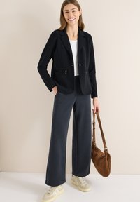 Zwarte blazer met twee ritssluitingen, gedragen over een wit shirt. Gecombineerd met grijze wijde broekspijpen en beige sneakers, met een bruine tas in de hand.